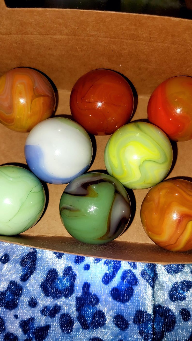 May include: A collection of colorful glass marbles in a cardboard box. The marbles feature swirls of orange, yellow, green, blue, and brown. The marbles are a classic toy, perfect for collectors or for playing games.