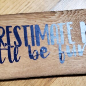May include: A rectangular wooden sign with the phrase "UNDERESTIMATE ME... that'll be fun..." in blue and light blue. The sign is made of light brown wood, showing the grain and knots.