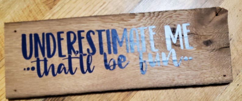 May include: A rectangular wooden sign with the phrase "UNDERESTIMATE ME... that'll be fun..." in blue and light blue. The sign is made of light brown wood, showing the grain and knots.