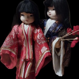 May include: Two Japanese dolls with black hair and traditional clothing. One doll wears a red kimono with gold accents, the other a blue and gold kimono. Both dolls have pale skin and are set against a black background.