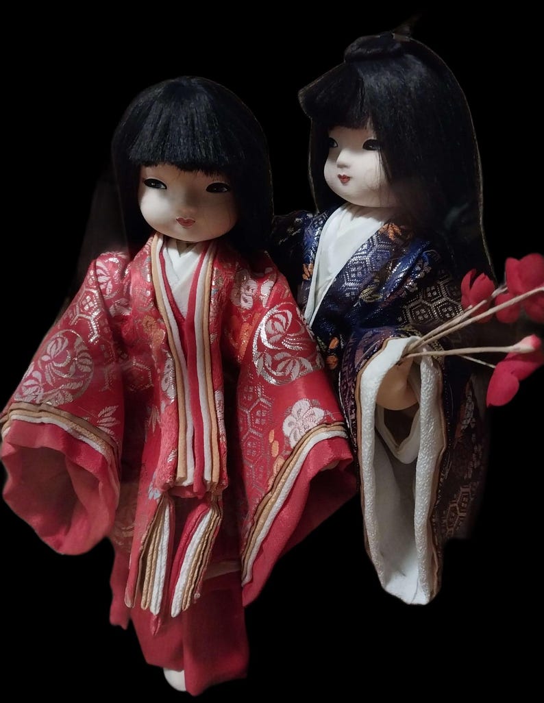 May include: Two Japanese dolls with black hair and traditional clothing. One doll wears a red kimono with gold accents, the other a blue and gold kimono. Both dolls have pale skin and are set against a black background.
