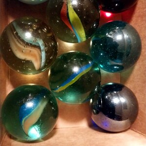 May include: A collection of colorful glass marbles in a cardboard box. The marbles vary in color, including blue, green, and black, with swirling patterns of yellow, red, and blue. Some marbles have a clear, transparent appearance.