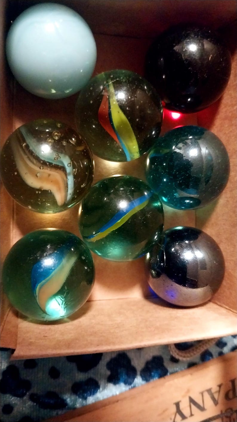 May include: A collection of colorful glass marbles in a cardboard box. The marbles vary in color, including blue, green, and black, with swirling patterns of yellow, red, and blue. Some marbles have a clear, transparent appearance.