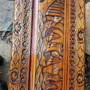 May include: A detailed close-up of a wooden chest lid with intricate carvings. The design features floral patterns along the edges and a central panel with a carved scene. The wood has a warm, natural tone.