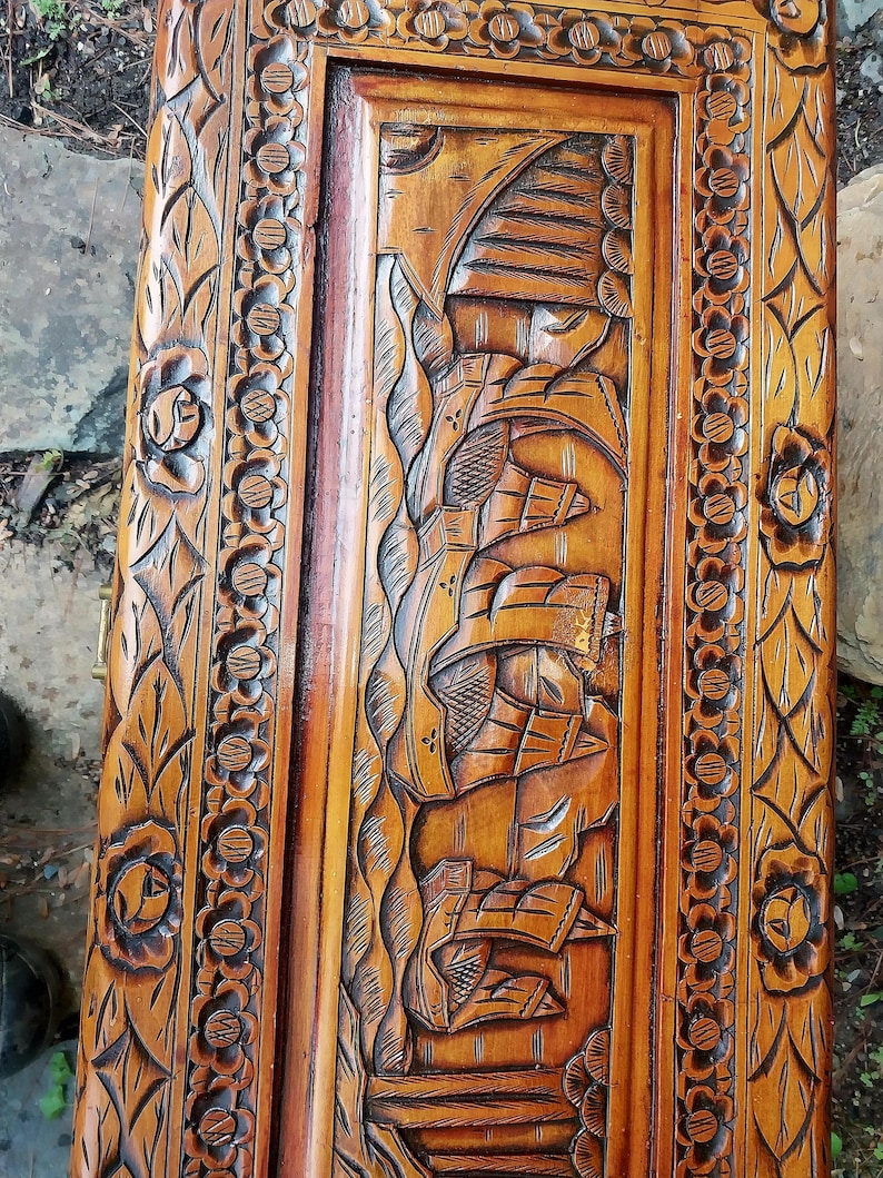 May include: A detailed close-up of a wooden chest lid with intricate carvings. The design features floral patterns along the edges and a central panel with a carved scene. The wood has a warm, natural tone.