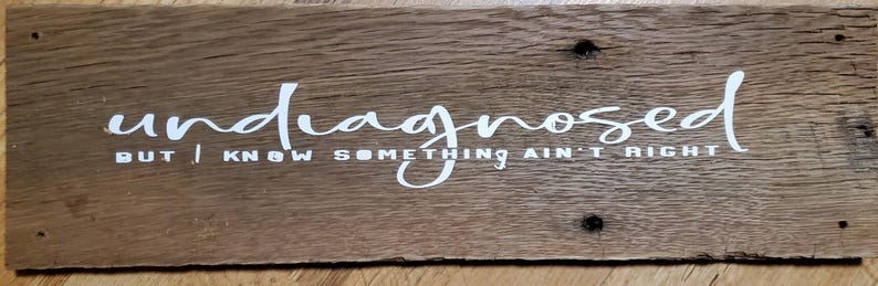 May include: A rectangular wooden sign with the words "undiagnosed BUT I KNOW SOMETHING AIN'T RIGHT" in white script. The sign is made of weathered wood with visible grain and knots, and has a rustic appearance.