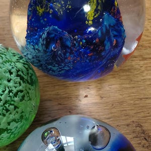 May include: Three glass paperweights. One is clear with a blue and yellow design, another is green, and the third is blue and clear with bubbles. The paperweights are on a wooden surface.