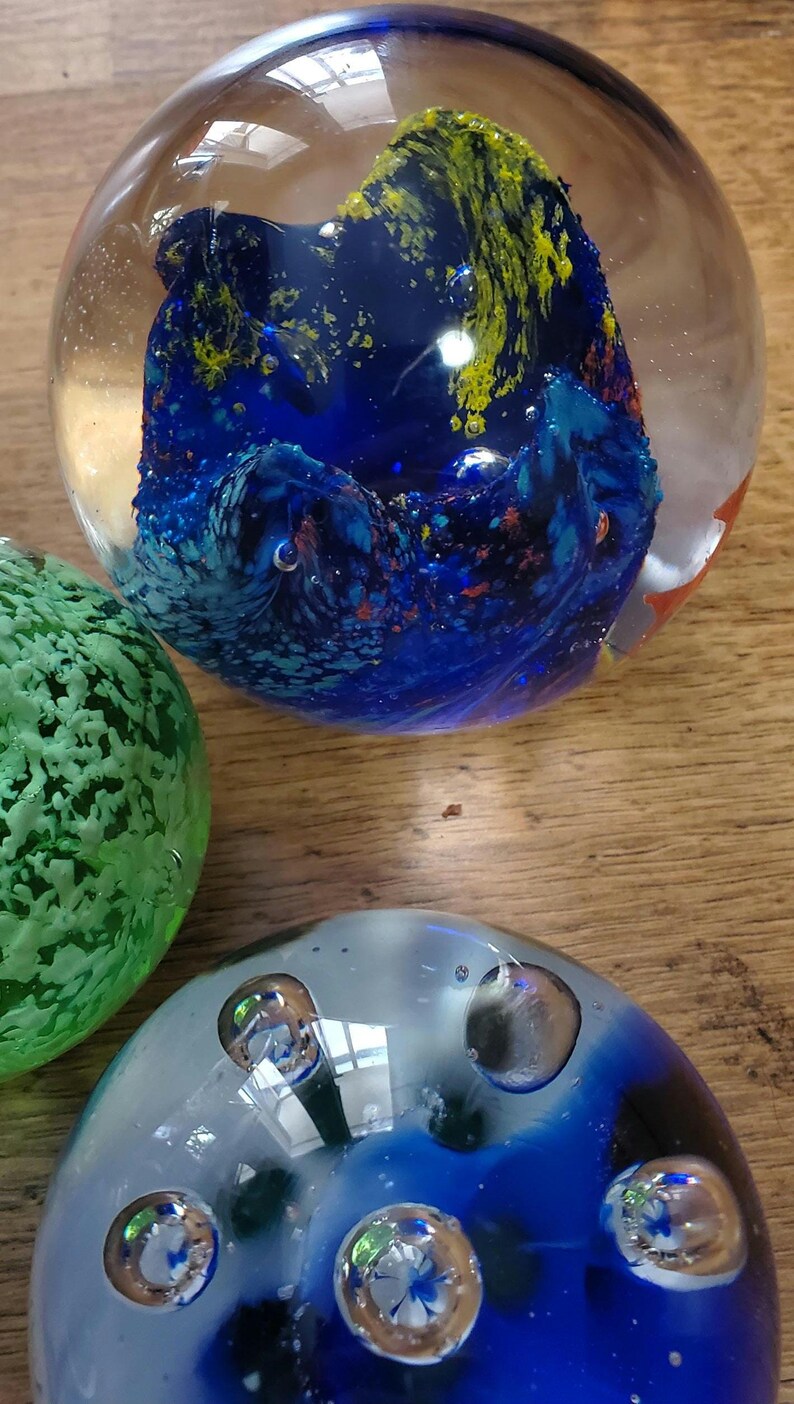 May include: Three glass paperweights. One is clear with a blue and yellow design, another is green, and the third is blue and clear with bubbles. The paperweights are on a wooden surface.