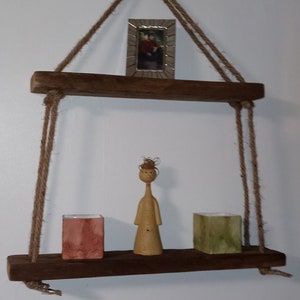 May include: A wooden shelf with rope hangers, featuring a small framed picture, a wooden figurine, and two small square containers. The shelf is hanging on a white wall.