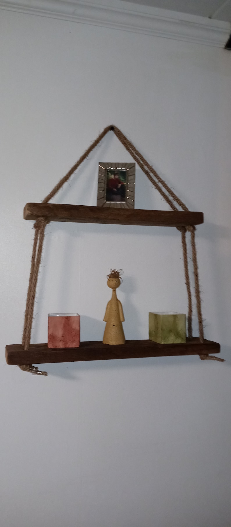 May include: A wooden shelf with rope hangers, featuring a small framed picture, a wooden figurine, and two small square containers. The shelf is hanging on a white wall.