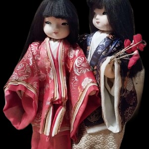 May include: Two Japanese dolls in traditional attire. The doll on the left wears a red kimono with gold accents, while the other doll wears a blue and gold kimono and holds red flowers. Both dolls have black hair and pale skin.