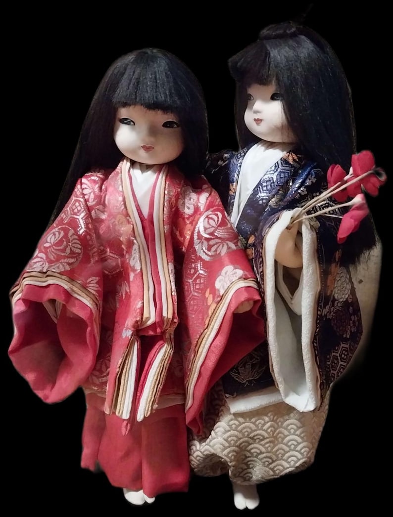 May include: Two Japanese dolls in traditional attire. The doll on the left wears a red kimono with gold accents, while the other doll wears a blue and gold kimono and holds red flowers. Both dolls have black hair and pale skin.