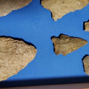 Pode incluir: Uma cole&ccedil;&atilde;o de antigas pontas de flecha e ferramentas de pedra, em v&aacute;rias formas e tamanhos, exibidas sobre um fundo azul. Os artefatos s&atilde;o principalmente em tons de bege e cinza, mostrando sua idade e artesanato.