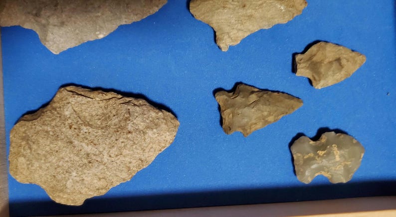 Pode incluir: Uma cole&ccedil;&atilde;o de antigas pontas de flecha e ferramentas de pedra, em v&aacute;rias formas e tamanhos, exibidas sobre um fundo azul. Os artefatos s&atilde;o principalmente em tons de bege e cinza, mostrando sua idade e artesanato.