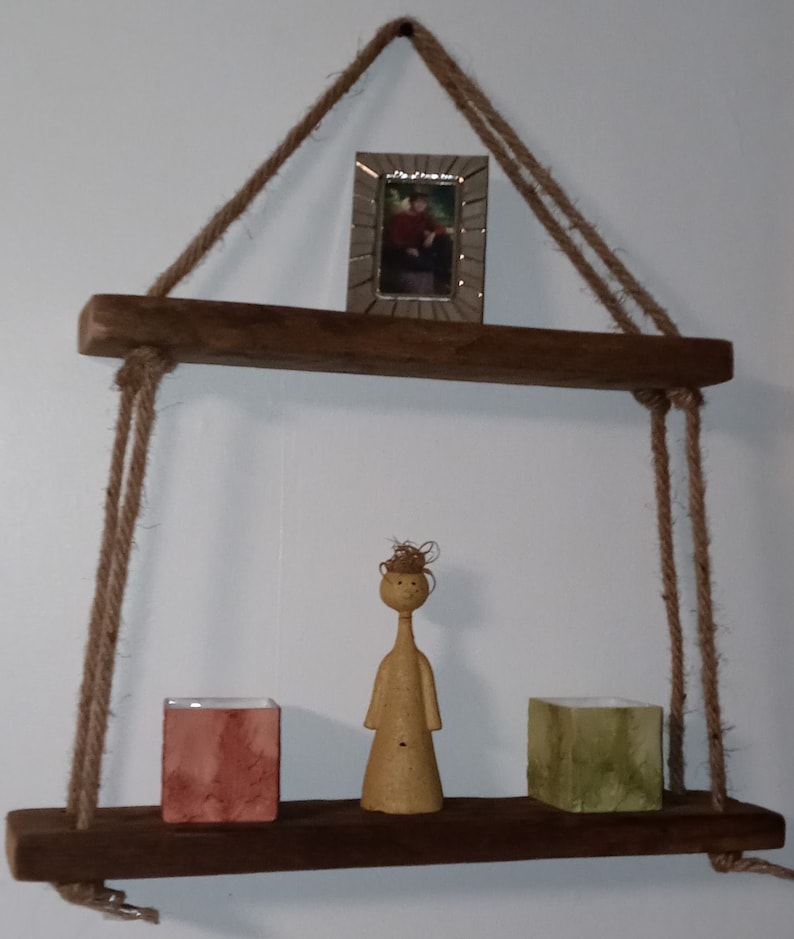 May include: A rustic wooden shelf with rope hangers, featuring a small framed picture, a ceramic figurine, and two square containers.
