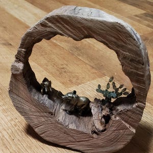 May include: A wooden sculpture featuring a circular frame with a carved-out interior. Inside, two small bronze elephant figurines are positioned near a small bronze tree. The wood has a natural, rough texture.