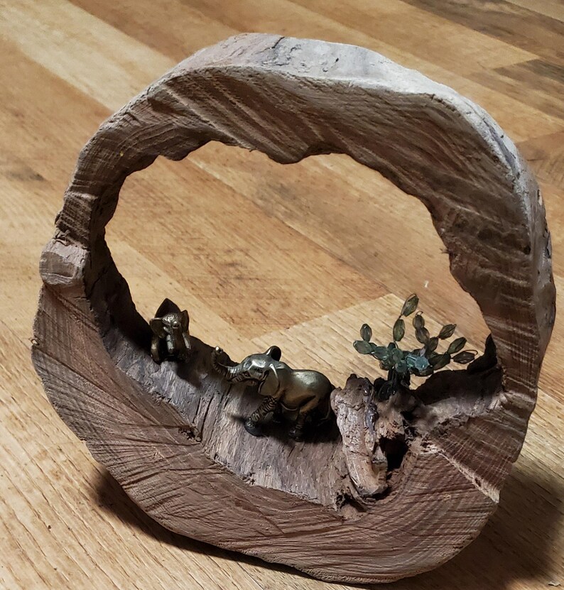 May include: A wooden sculpture featuring a circular frame with a carved-out interior. Inside, two small bronze elephant figurines are positioned near a small bronze tree. The wood has a natural, rough texture.