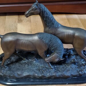 May include: Bronze sculpture of two horses. One horse stands tall, while the other lowers its head. The horses are on a dark, textured base. The sculpture has a dark brown patina and is a decorative item.