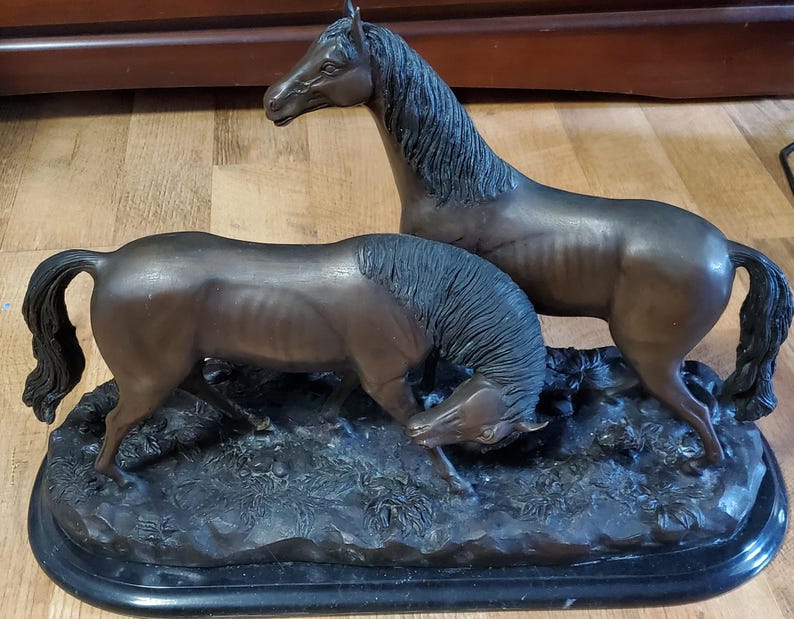 May include: Bronze sculpture of two horses. One horse stands tall, while the other lowers its head. The horses are on a dark, textured base. The sculpture has a dark brown patina and is a decorative item.