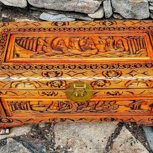 May include: A rectangular wooden chest with intricate carvings. The chest features a detailed relief of sailing ships on the lid and floral patterns on the sides. It has a brass clasp and is stained in a warm, reddish-brown hue.