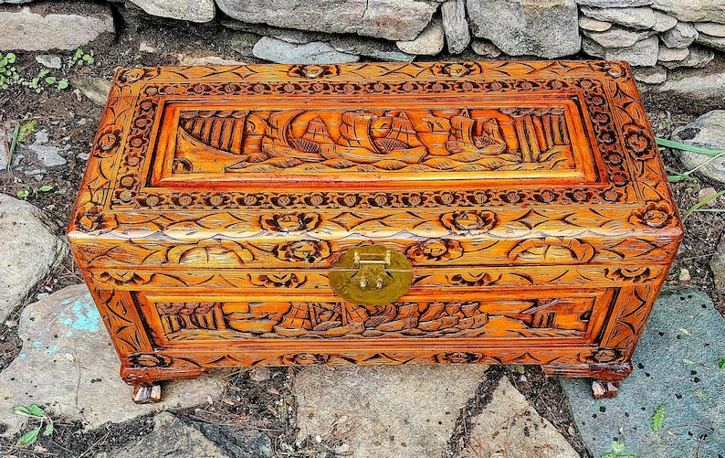 May include: A rectangular wooden chest with intricate carvings. The chest features a detailed relief of sailing ships on the lid and floral patterns on the sides. It has a brass clasp and is stained in a warm, reddish-brown hue.