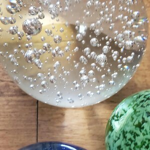 May include: A selection of decorative glass paperweights. The largest is clear with many silver bubbles inside. A green and a blue paperweight are also visible, each with unique patterns and colours. The objects are on a wooden surface.