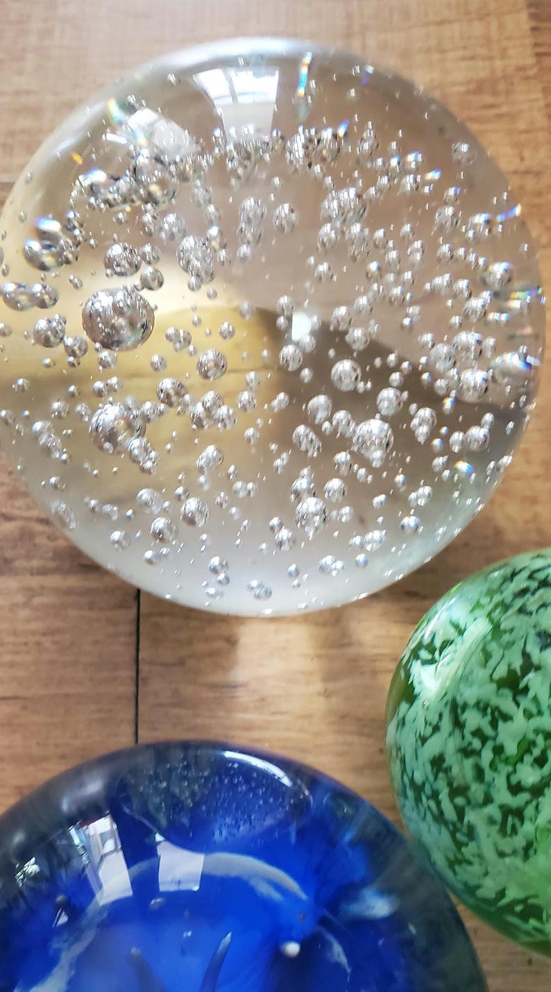 May include: A selection of decorative glass paperweights. The largest is clear with many silver bubbles inside. A green and a blue paperweight are also visible, each with unique patterns and colours. The objects are on a wooden surface.