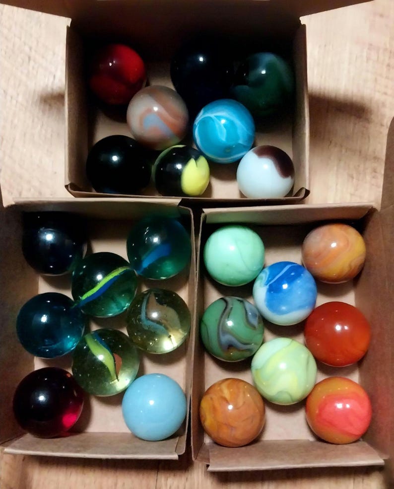 May include: A collection of colorful glass marbles in a cardboard box. The marbles are various colors, including red, blue, green, yellow, and orange, with swirling patterns and solid colors. Some marbles have a translucent appearance.