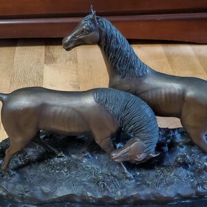 May include: Bronze sculpture of two horses. One horse stands tall, the other is grazing. The horses have detailed manes and tails, and are set on a dark, textured base. The sculpture is a decorative item.
