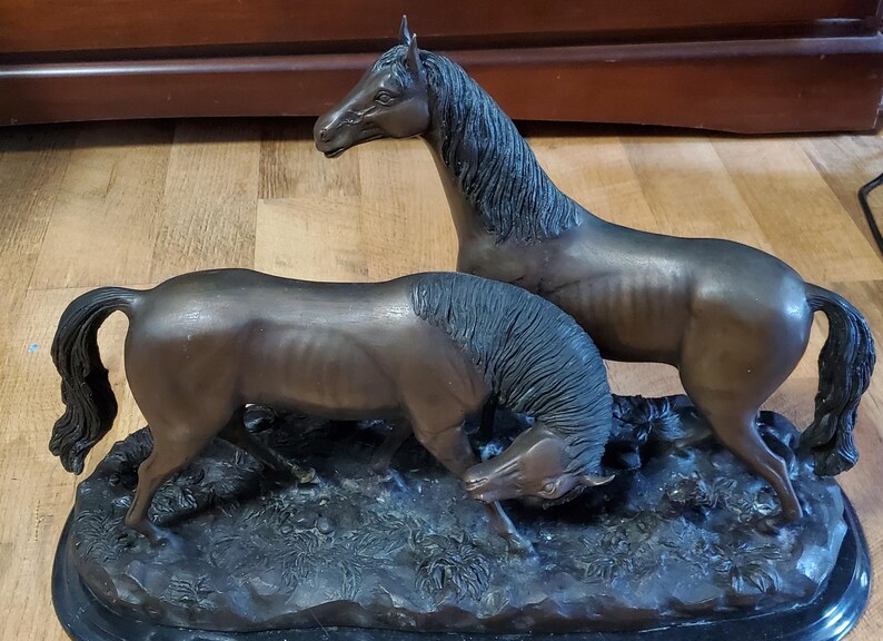 May include: Bronze sculpture of two horses. One horse stands tall, the other is grazing. The horses have detailed manes and tails, and are set on a dark, textured base. The sculpture is a decorative item.