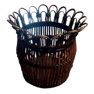 May include: A dark brown woven basket with a cylindrical body and a decorative scalloped rim. The structure is composed of vertical reed or bamboo slats held together by horizontal bands, creating an open, airy design with a circular base.