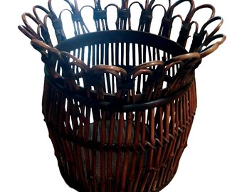 Puede incluir: Una cesta trenzada de color marrón oscuro con cuerpo cilíndrico y un borde festoneado decorativo. La estructura está compuesta por listones verticales de caña o bambú unidos por bandas horizontales, creando un diseño abierto y circular.