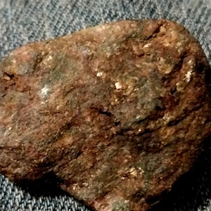 May include: A close-up of a rough, irregularly shaped rock. The rock has a reddish-brown color with patches of green and small, shiny, white flecks. The surface appears textured and weathered, suggesting exposure to the elements.