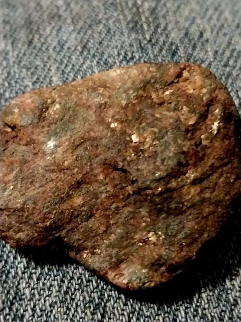 May include: A close-up of a rough, irregularly shaped rock. The rock has a reddish-brown color with patches of green and small, shiny, white flecks. The surface appears textured and weathered, suggesting exposure to the elements.