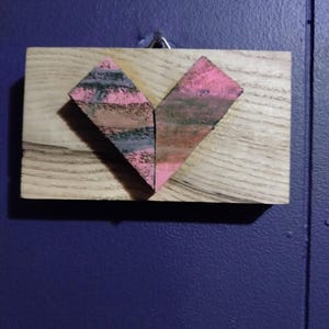 May include: A wooden plaque with a heart-shaped design made of pink and gray textured paper. The heart is centered on the plaque.