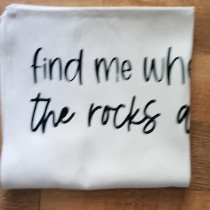 May include: White folded cloth with the text "find me where the rocks are" in black cursive font. The cloth is rectangular and appears to be made of a soft material. The background is a wooden surface.