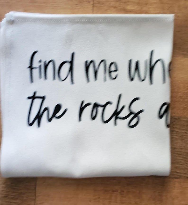 May include: White folded cloth with the text "find me where the rocks are" in black cursive font. The cloth is rectangular and appears to be made of a soft material. The background is a wooden surface.