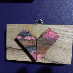 May include: A wooden heart-shaped plaque with a pink and black painted design. The plaque is attached to a wall with a metal hook.