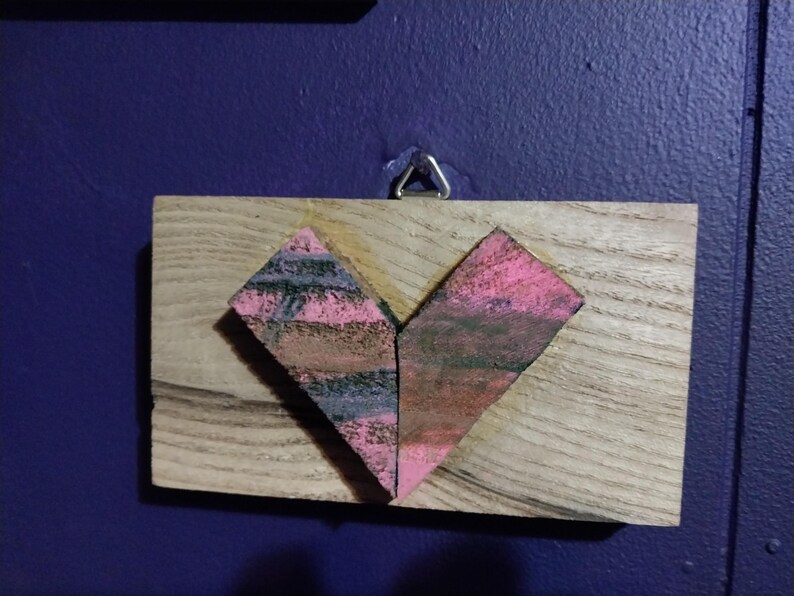 May include: A wooden heart-shaped plaque with a pink and black painted design. The plaque is attached to a wall with a metal hook.