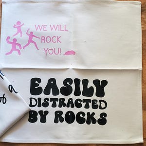 May include: Off-white cotton tea towel with pink text and graphics. The top half reads "WE WILL ROCK YOU!" with three stick figures. The bottom half reads "EASILY DISTRACTED BY ROCKS" in black, bold, bubble letters.