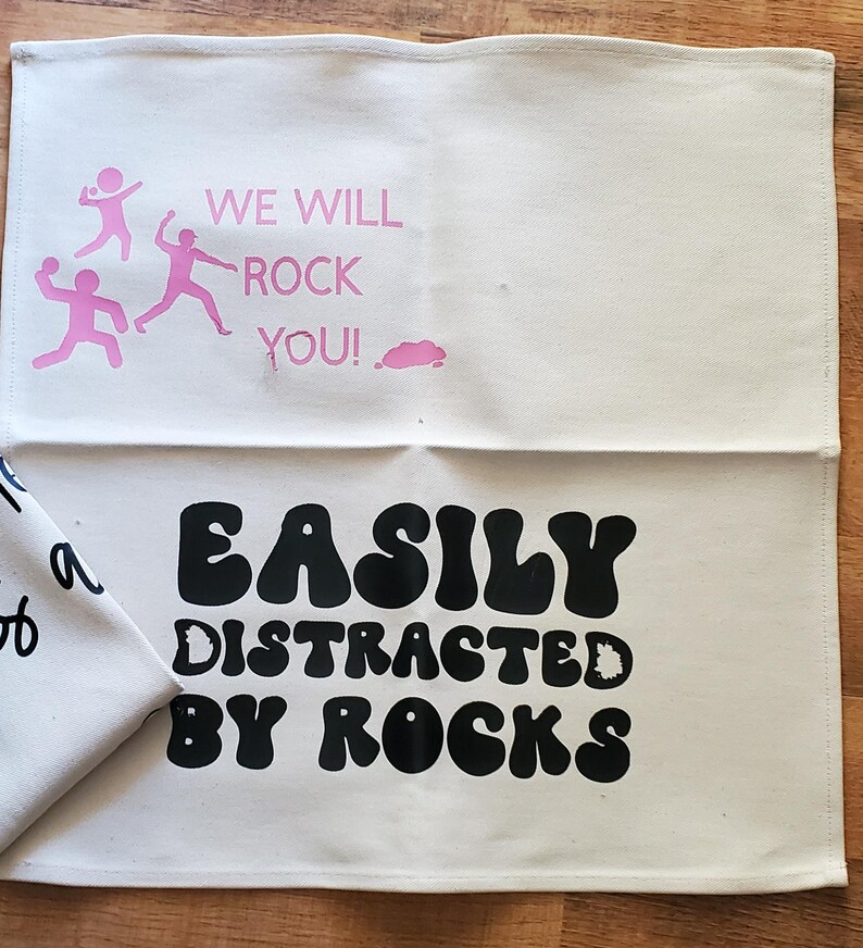 May include: Off-white cotton tea towel with pink text and graphics. The top half reads "WE WILL ROCK YOU!" with three stick figures. The bottom half reads "EASILY DISTRACTED BY ROCKS" in black, bold, bubble letters.