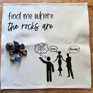 May include: A white cloth with the text "find me where the rocks are" and a cartoon illustration. The illustration includes a person pointing at a group of rocks, speech bubbles, and silhouettes of people. Various rocks are displayed.