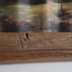 May include: A framed landscape print featuring a bridge over a stream in a wooded area. The frame is made of rustic, light brown wood. The artwork depicts a serene outdoor scene with trees and foliage.