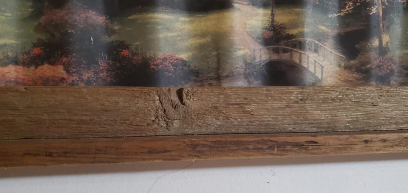 May include: A framed landscape print featuring a bridge over a stream in a wooded area. The frame is made of rustic, light brown wood. The artwork depicts a serene outdoor scene with trees and foliage.