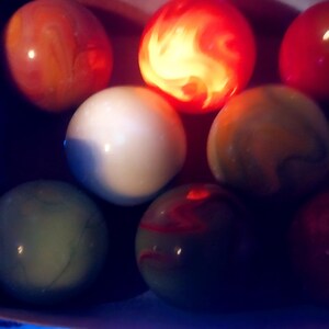 May include: A collection of colorful marbles in various shades of red, orange, white, green, and yellow. Some marbles have swirling patterns, and one is illuminated with a bright, fiery glow. The marbles are arranged in a dark setting.