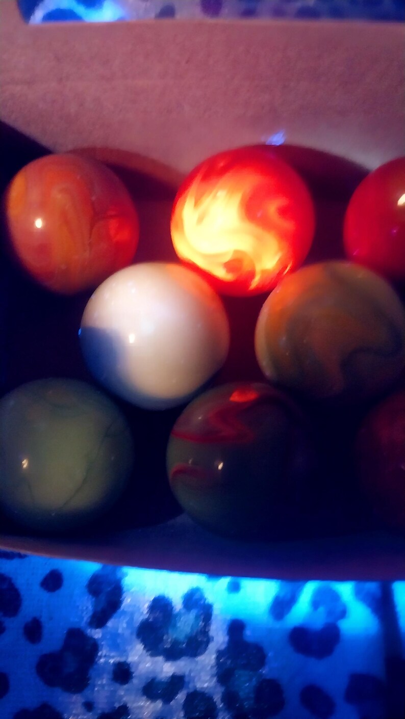 May include: A collection of colorful marbles in various shades of red, orange, white, green, and yellow. Some marbles have swirling patterns, and one is illuminated with a bright, fiery glow. The marbles are arranged in a dark setting.