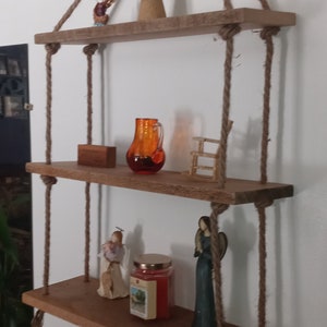 May include: Three wooden shelves hanging from the ceiling with rope. The shelves are decorated with small figurines, a glass pitcher, and a candle.