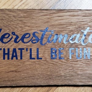 May include: A rectangular wooden sign with the words "Underestimate me, that'll be fun" in blue and silver lettering. The sign has a rustic, weathered appearance with visible wood grain and screw holes.