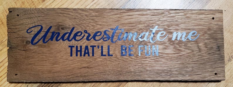 May include: A rectangular wooden sign with the words "Underestimate me, that'll be fun" in blue and silver lettering. The sign has a rustic, weathered appearance with visible wood grain and screw holes.