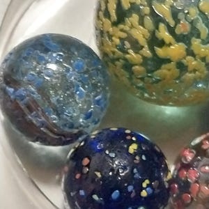 Op de afbeelding: Een verzameling van vier glazen knikkers met verschillende patronen. E&eacute;n knikker is blauw met witte stippen, een andere is groen met gele stippen, een andere is blauw met veelkleurige stippen en de laatste is helder met rode en witte stippen.